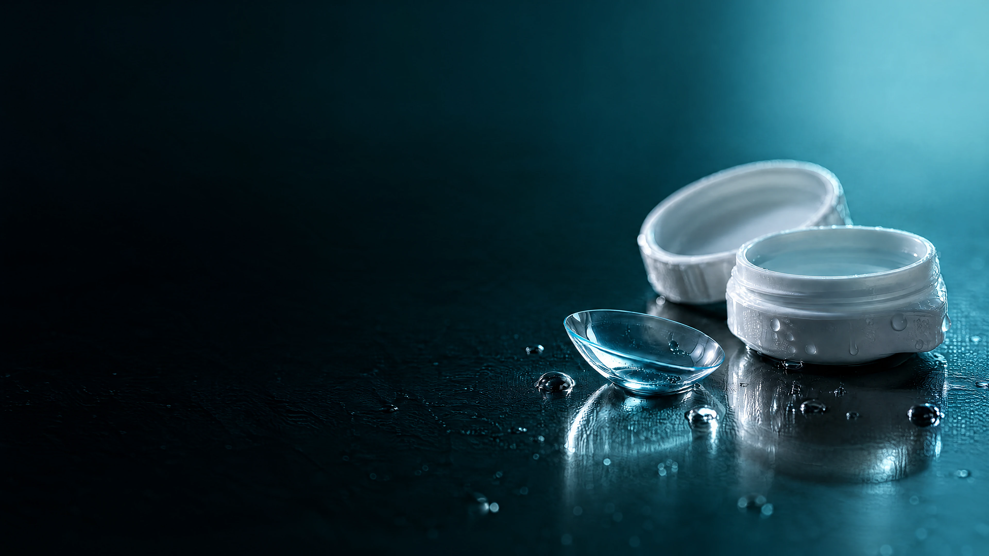 A soft contact lens beside an open lens case, with water droplets on a dark surface