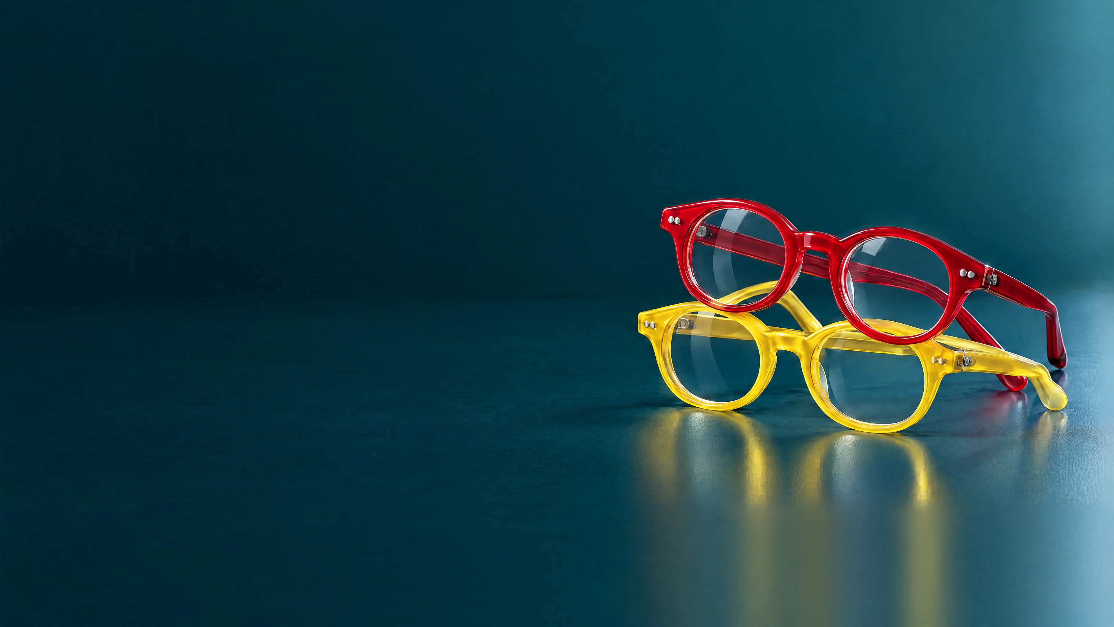 Two pairs of children's eyeglasses, one red and one yellow, stacked on a teal surface
