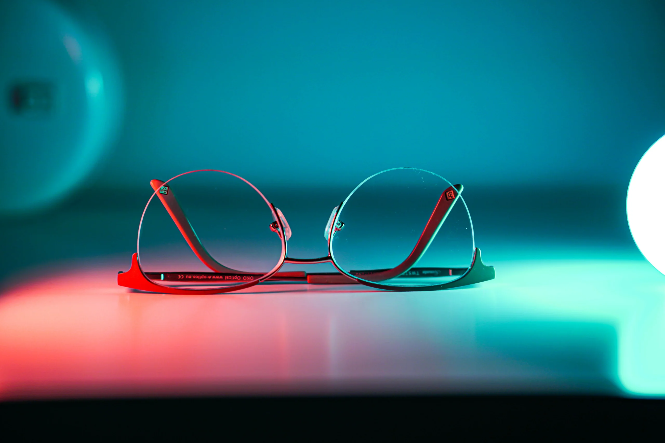 A pair of designer eyeglass frames resting on a lit surface under teal and red studio light