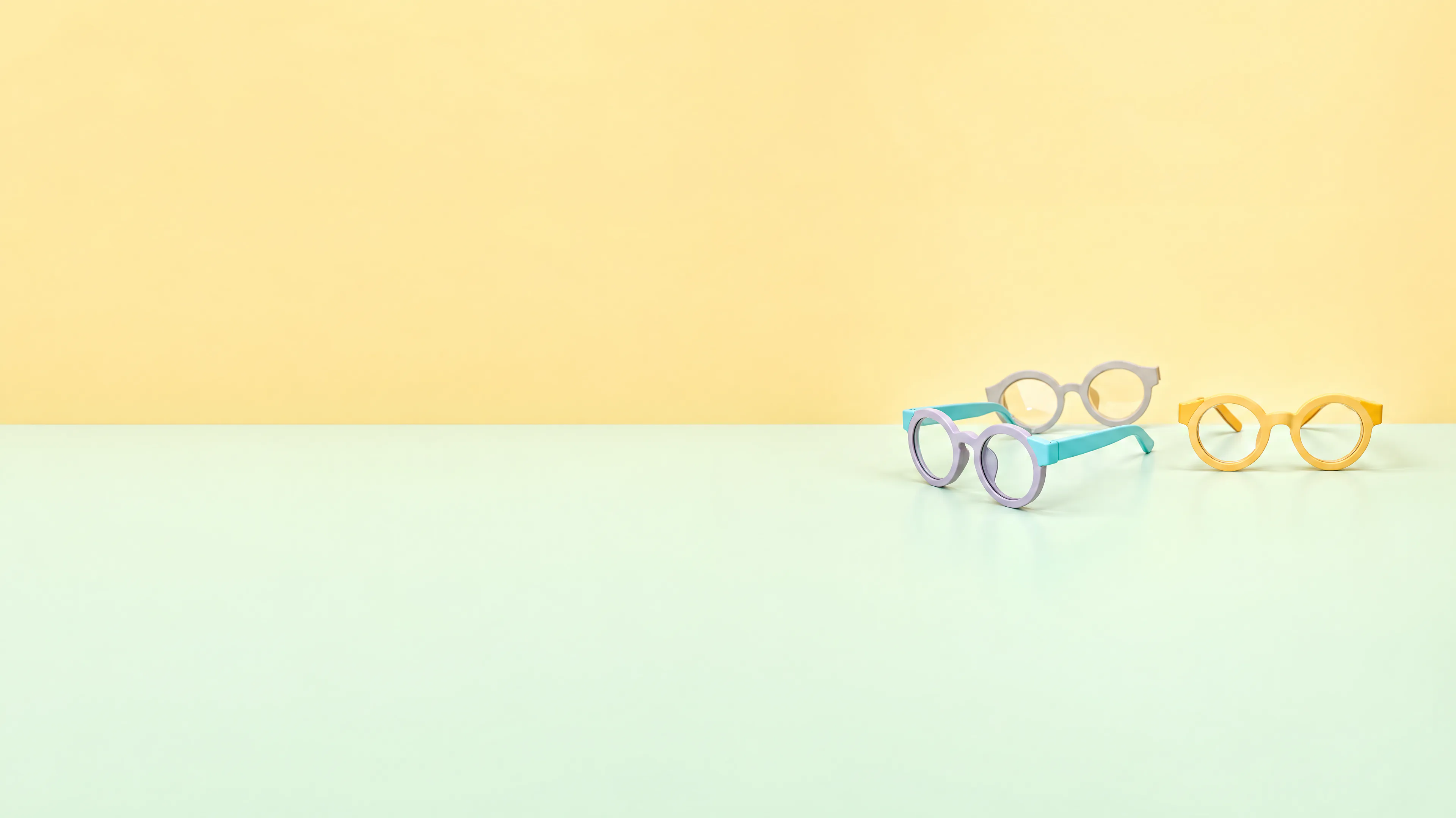 Three pairs of round children's eyeglass frames in lilac with teal arms, grey, and yellow, on a pale mint and yellow studio backdrop