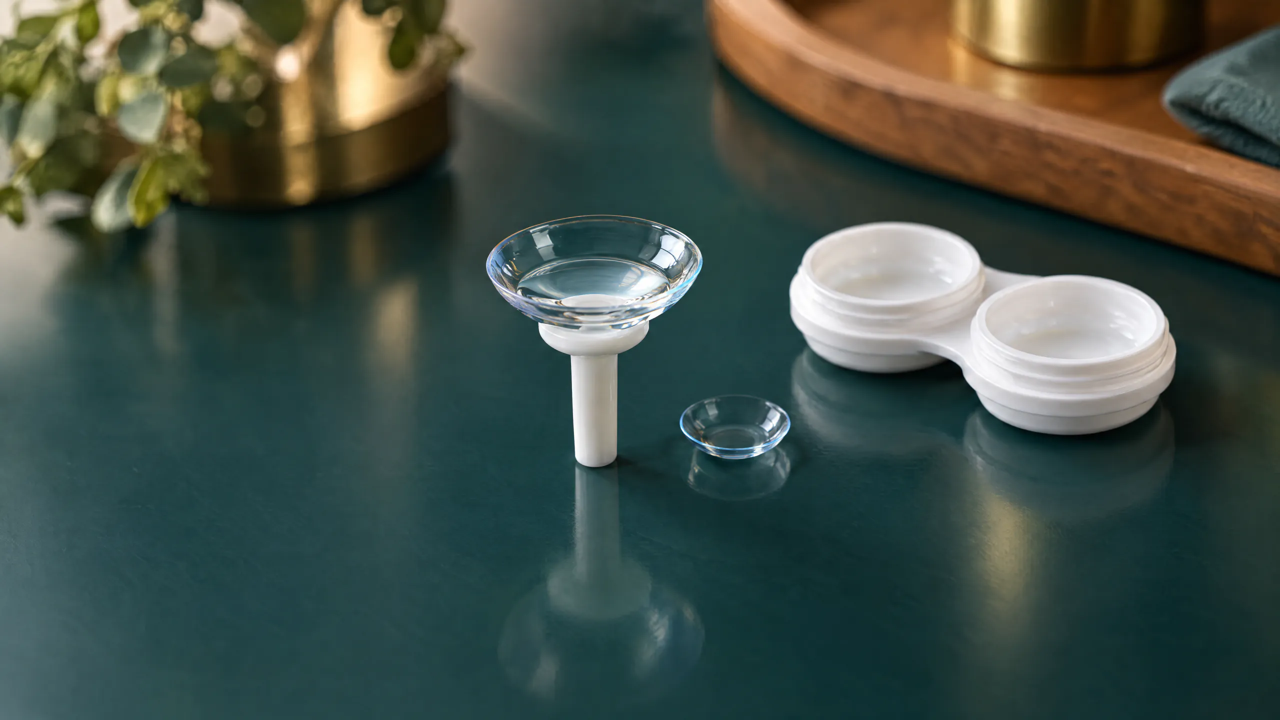 A large clear scleral contact lens on a white plunger beside a much smaller soft contact lens and an open white lens case, on a glossy dark teal surface