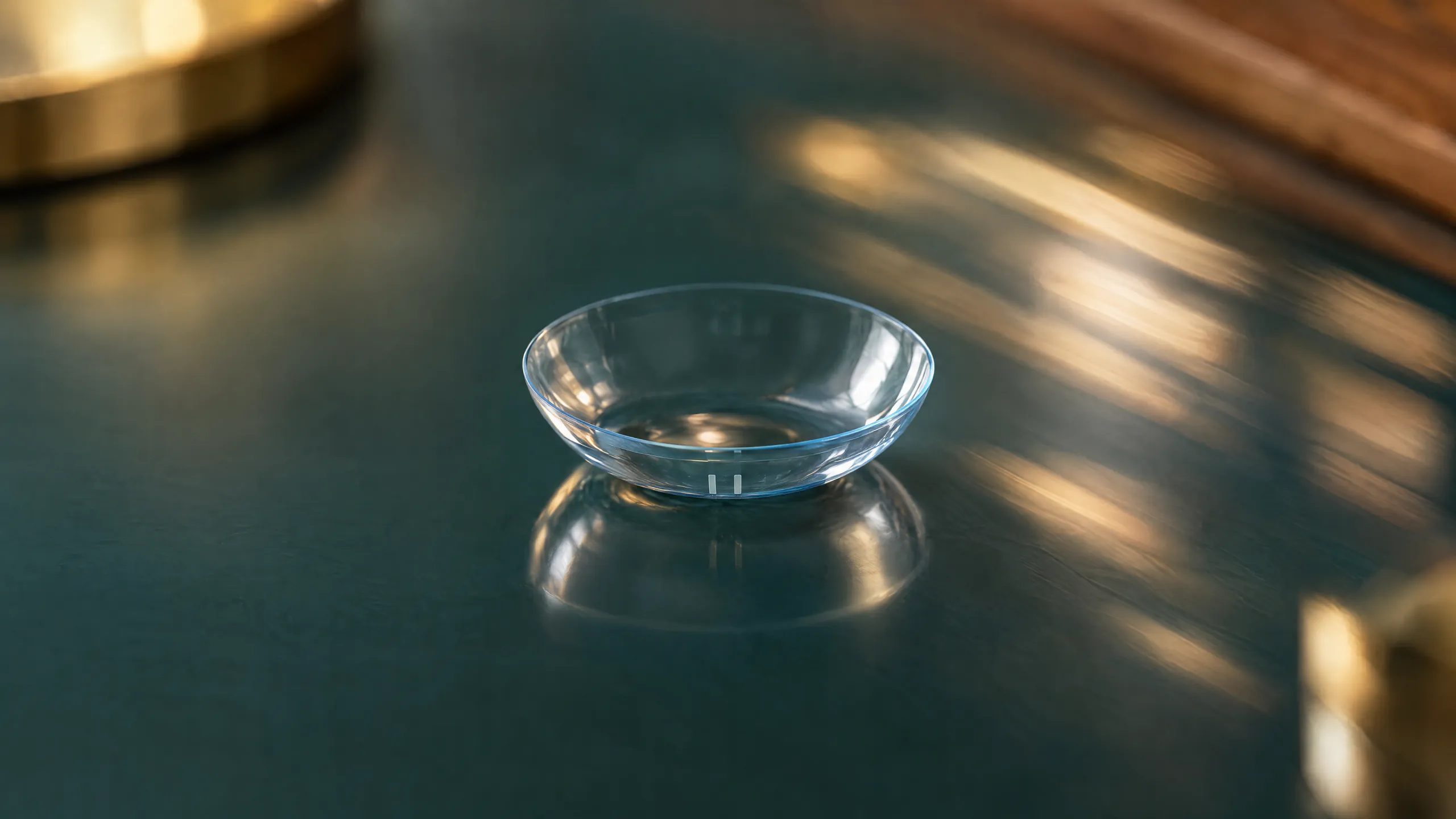 A clear soft contact lens with a thin blue edge and two small orientation marks, on a glossy dark teal surface crossed by streaks of light